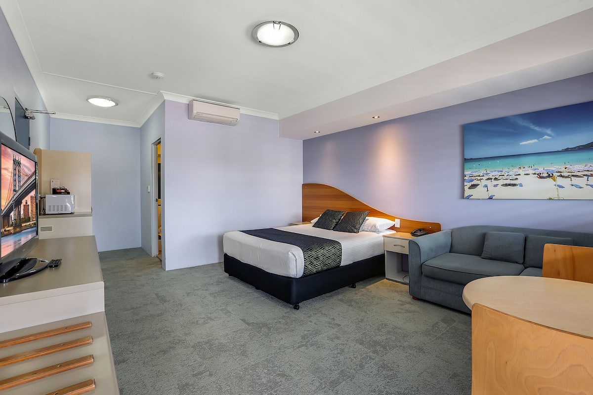 Habitación de hotel con cama, escritorio, silla, televisión y un cuadro de una playa.