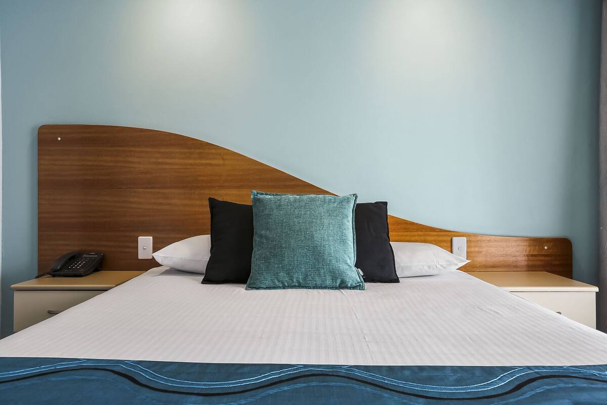 Una cama bien tendida con un edredón blanco, una almohada de color turquesa y dos almohadas negras. Se aprecia un cabecero de madera, una mesita de noche con un teléfono y un interruptor de pared.
