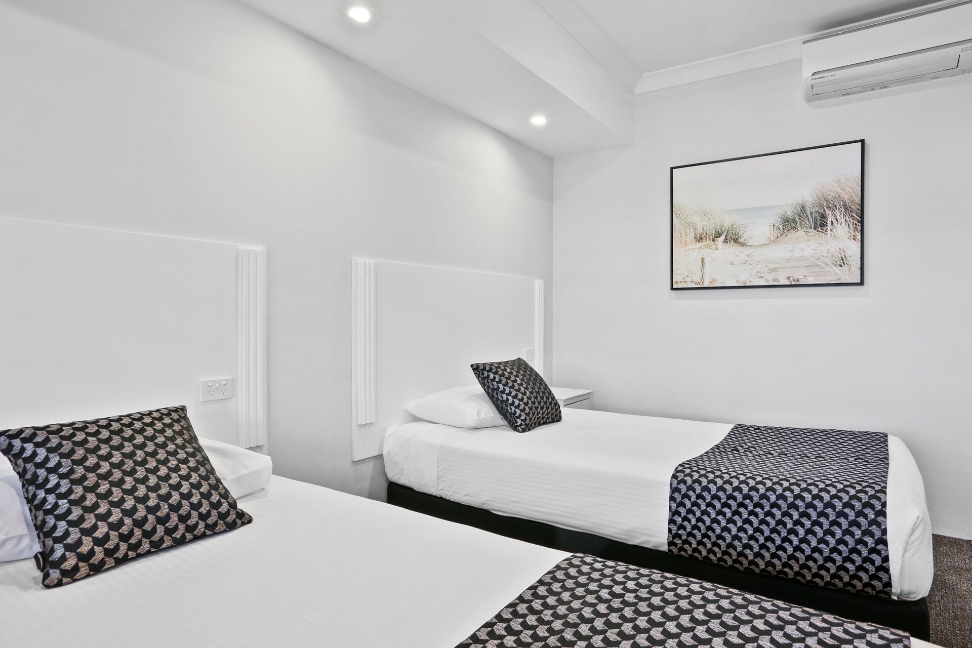 Habitación de hotel con dos camas individuales, ropa de cama blanca, almohadas estampadas, un cuadro en la pared y un equipo de aire acondicionado.