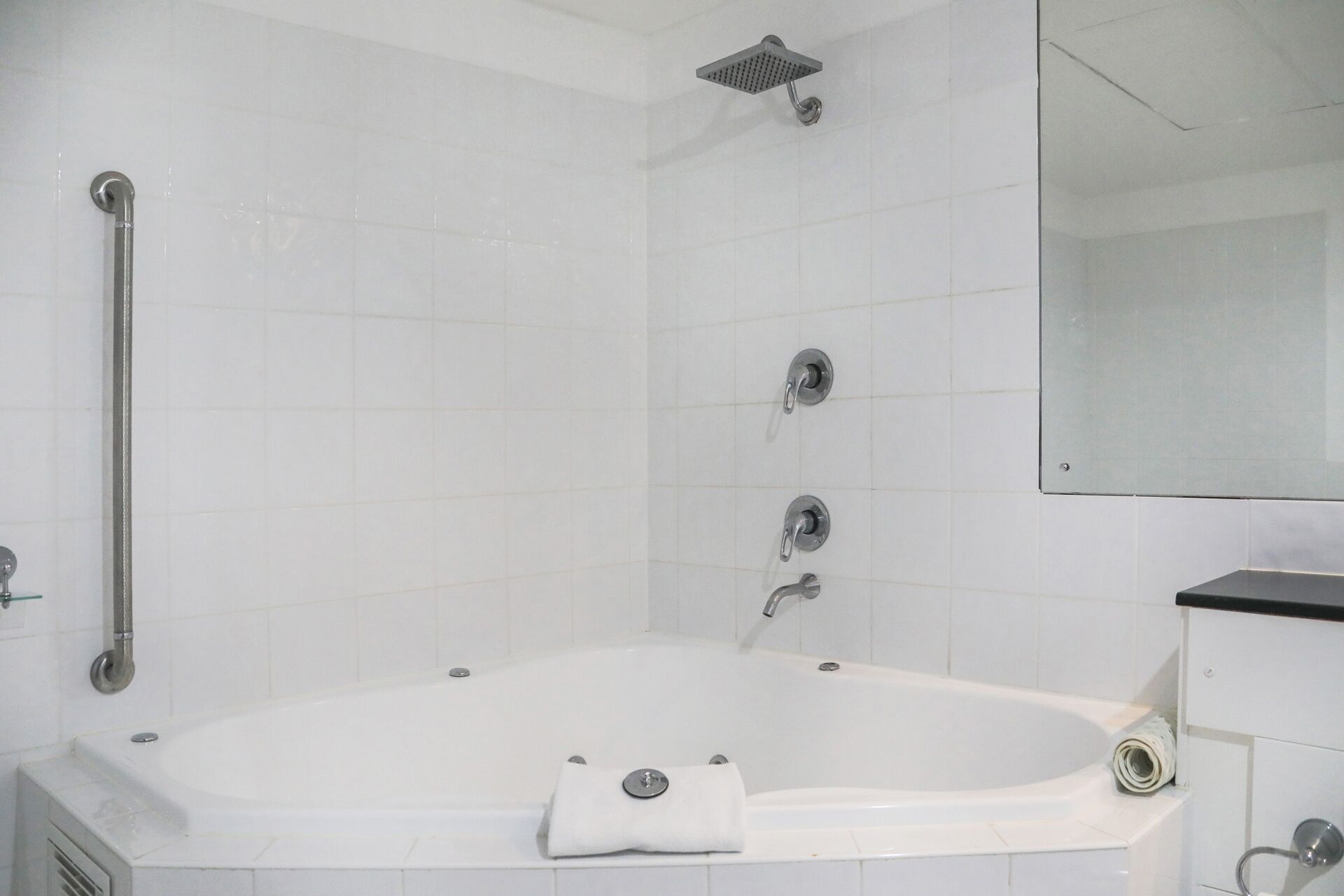 A bathroom with a white bathtub, a showerhead, a handrail, and a mirror.