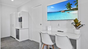 Une chambre d’hôtel moderne avec un bureau blanc, deux chaises blanches, un téléviseur à écran plat fixé au mur, une petite plante en pot et un mini-bar.