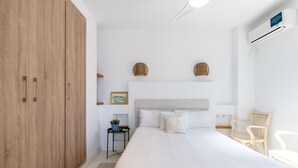 A bedroom with a large wooden wardrobe, a bed with white linens, a small side table with a plant, and a wicker chair. - Alfarería Terrace. 2 bedrooms, private terrace (Sevilla)