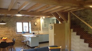 Un intérieur chaleureux avec un mur en pierre, des poutres en bois, un coin repas avec une table et des chaises, un canapé avec des coussins à rayures et un escalier menant à un niveau supérieur.