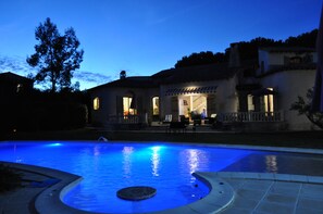 Una piscina con un jacuzzi circular, un patio con mobiliario exterior, y una casa con tejado de tejas y varias ventanas.