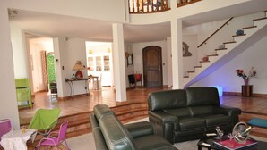 A spacious living room with a black leather sofa, a green chair, and a dining table with chairs. A staircase leads to an upper level. - Family home, swimming pool, 5 bedrooms with private bathrooms Enclosed garden (Saint-Geniès-De-Malgoirès)