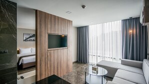 Ein modernes Hotelzimmer mit einem Bett, einem Sofa, einem Glastisch, einem Fernseher und einem großen Fenster mit Vorhängen.