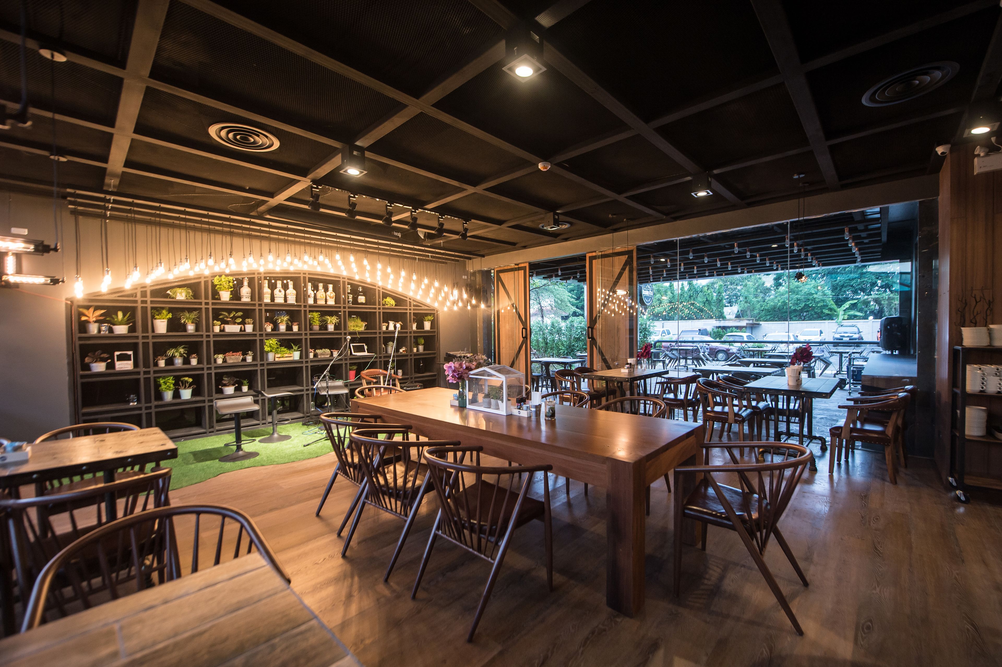 Un restaurant moderne avec des tables et des chaises en bois, un bar central agrémenté d’étagères remplies de bouteilles et de plantes, et de grandes fenêtres offrant une vue sur l’extérieur.