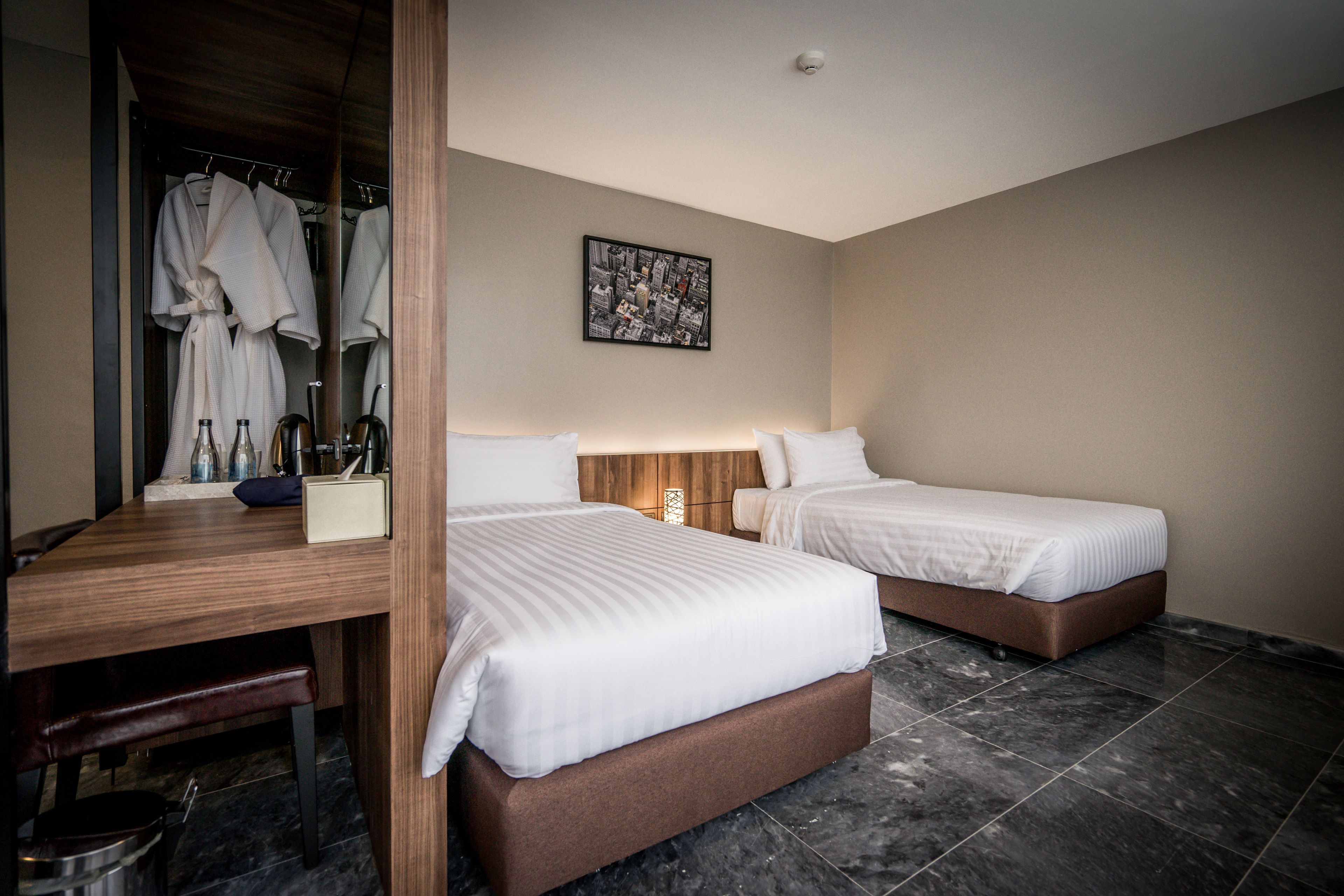 A hotel room with two beds, a wooden desk, a mirror, and a cityscape artwork on the wall.