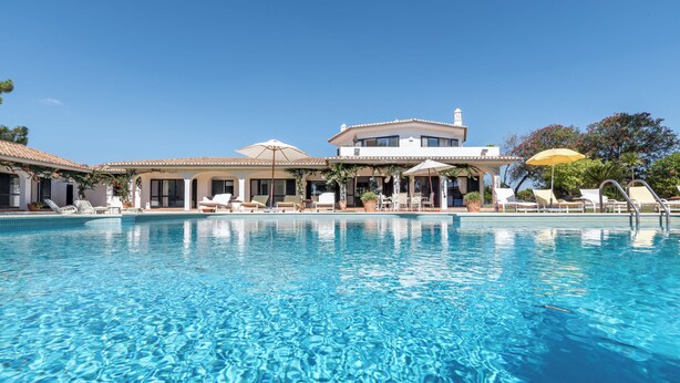 Une piscine d’un bleu éclatant, bordée de chaises longues et d’ombrelles, et entourée d’un bâtiment doté d’un toit en tuiles et de grandes fenêtres.