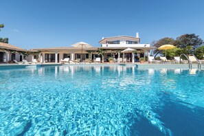 Une piscine d’un bleu éclatant, bordée de chaises longues et d’ombrelles, et entourée d’un bâtiment doté d’un toit en tuiles et de grandes fenêtres.