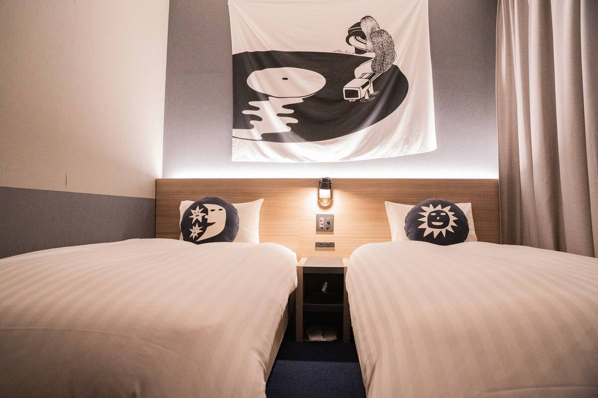 A hotel room with two beds, a nightstand, a wall-mounted lamp, and a tapestry with a graphic design.