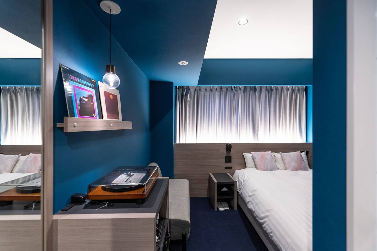 A hotel room with a bed, bedside table, a desk with a laptop, a mirror, and a window with curtains.