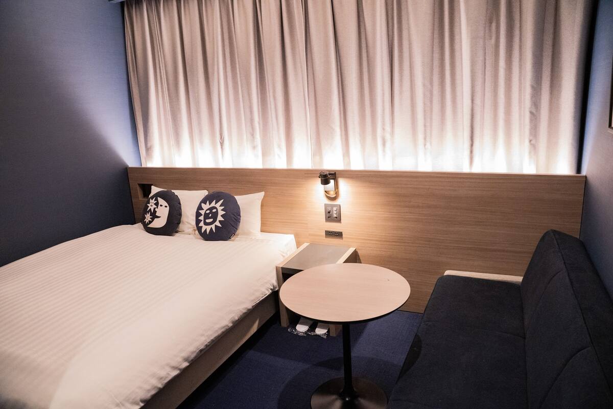 A hotel room with a bed, a small round table, a sofa, and a nightstand with a lamp.
