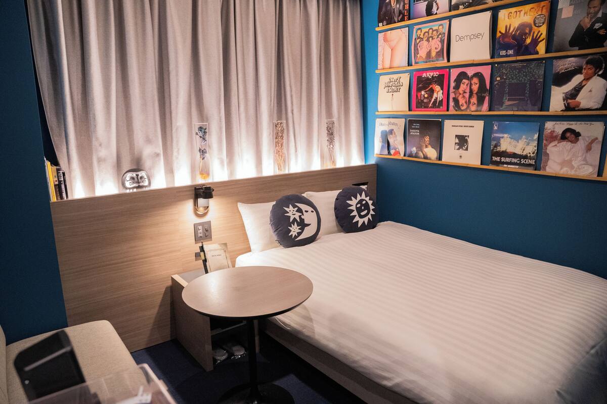 A hotel room with a bed, a small table, a nightstand, and a wall decorated with album covers.
