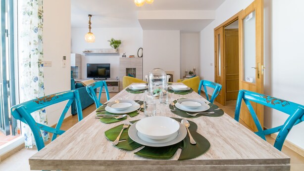 A dining area with a wooden table set for six, surrounded by blue chairs, and a kitchen in the background. - Parque Tecnológico Villa, ideal for couples and families. Swimming pool, games, park, etc. (Málaga)