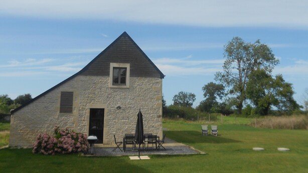 A stone building with a triangular roof, a door, and a window. There are outdoor chairs and a table with a table umbrella.