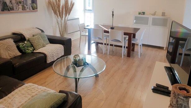 Een moderne woonkamer met een glazen salontafel, een zwarte bank en een eethoek met witte stoelen.