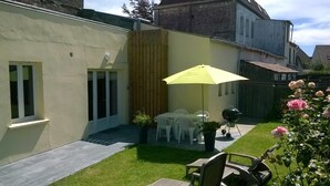 Een terras met een gele parasol, witte plastic stoelen, een barbecue en een kleine rozen tuin.