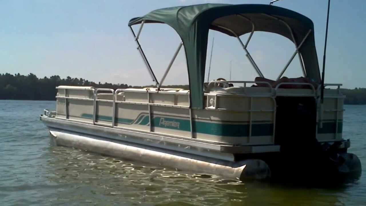 Een pontonboot met een zonnedak, voorzien van zitplaatsen en een klein hok, drijvend op een rustig wateroppervlak.