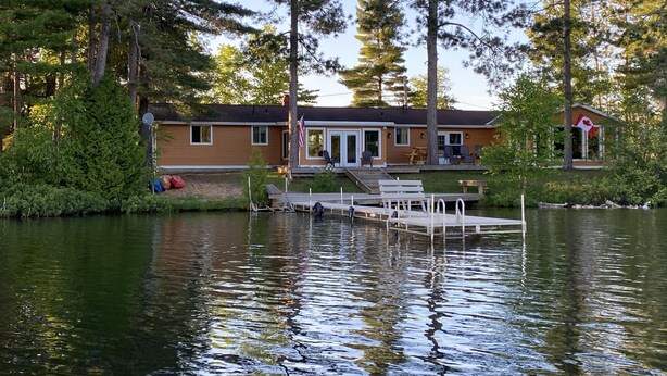 Un chalet au bord du lac, doté d’un quai, entouré de grands arbres et arborant un drapeau canadien.