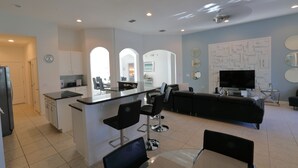 A modern kitchen with a central island, bar stools, and a dining area with a TV and seating. - Welcome To Our Beautiful Villa In The Gorgeous Community Solterra! (Davenport)