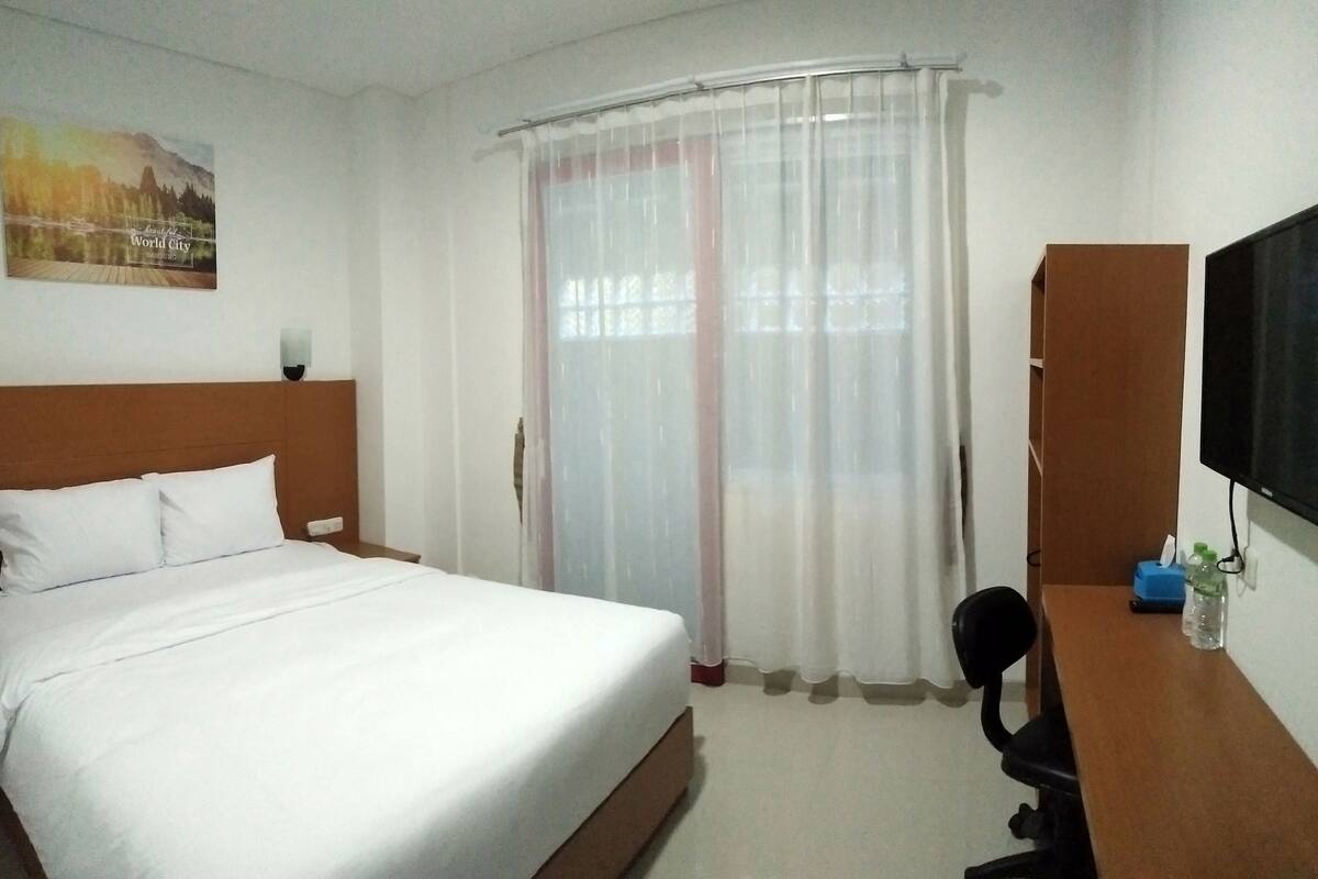 A hotel room with a bed, a desk, a chair, a television, and a window with sheer curtains.