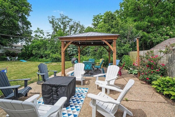 A backyard patio with Adirondack chairs, a covered seating area, and a checkered outdoor rug.