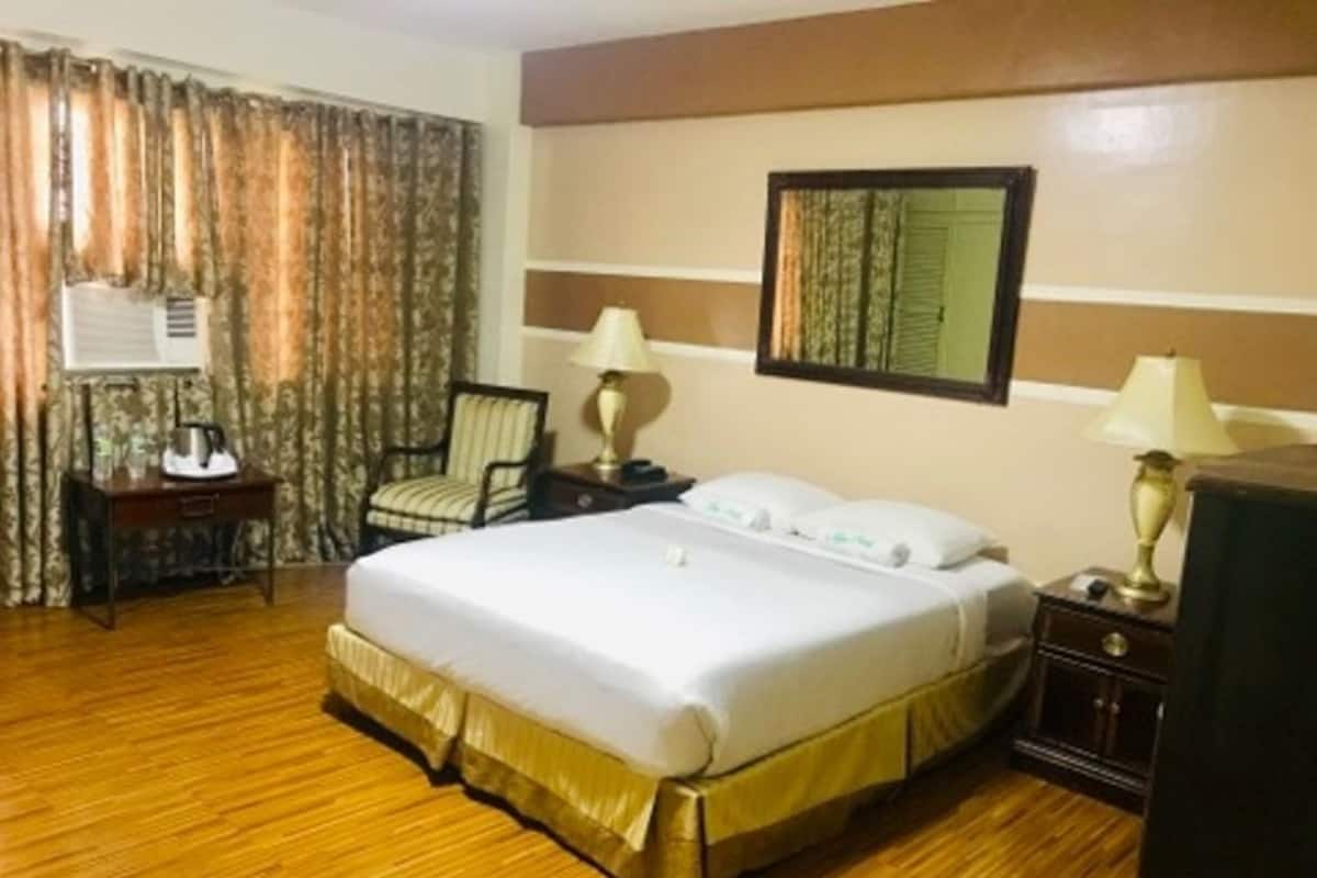 Hotelkamer met een groot bed, twee nachtkastjes met lampen, een stoel, een bureau met telefoon en een raam met gordijnen.