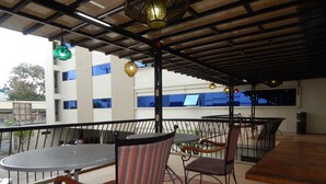 Un espacio de estar al aire libre cubierto con mesas y sillas de metal, una barandilla y luces colgantes.