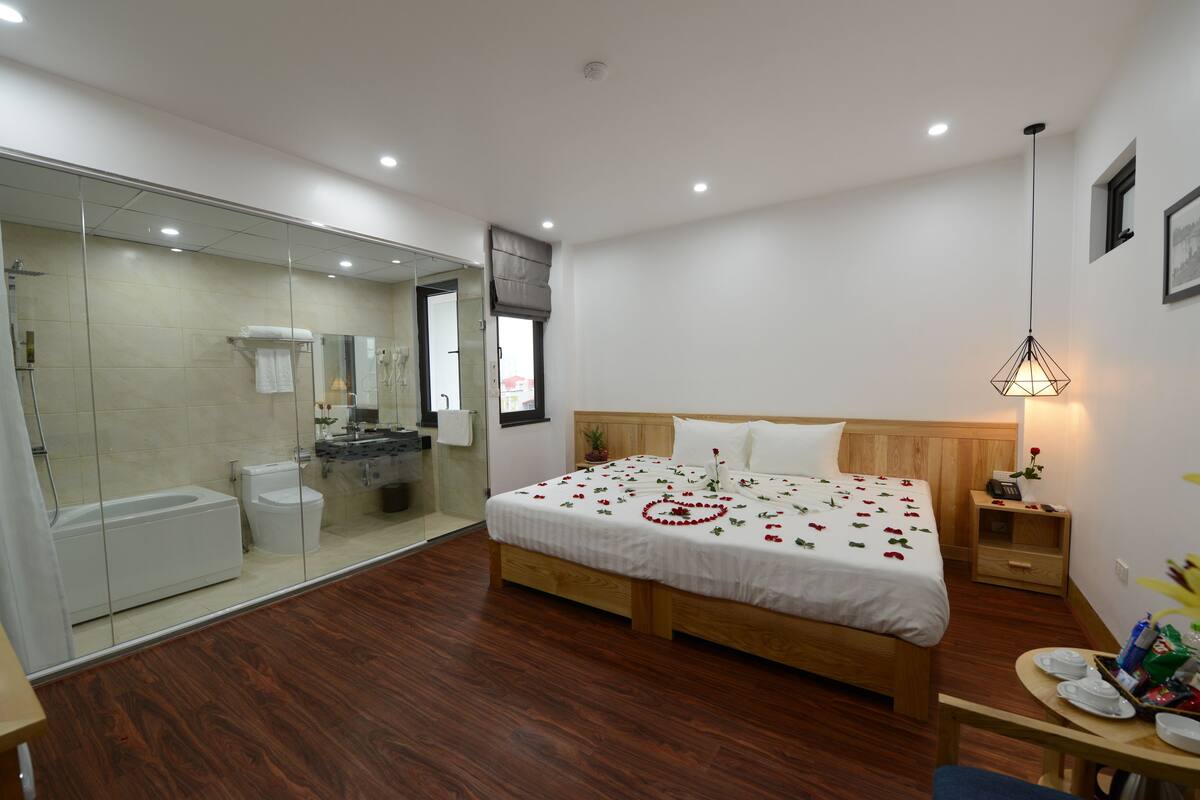 A modern hotel room with a wooden bed, a glass shower enclosure, a bedside table with a lamp, and a small table with a vase of flowers.