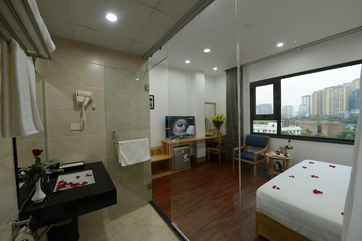 A hotel room with a bed, a desk, a chair, a small table with a vase, a mirror, and a view of the city through the window.