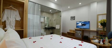 Una habitación de hotel con una cama grande, televisión, escritorio con computadora y baño visible a través de una puerta de vidrio.