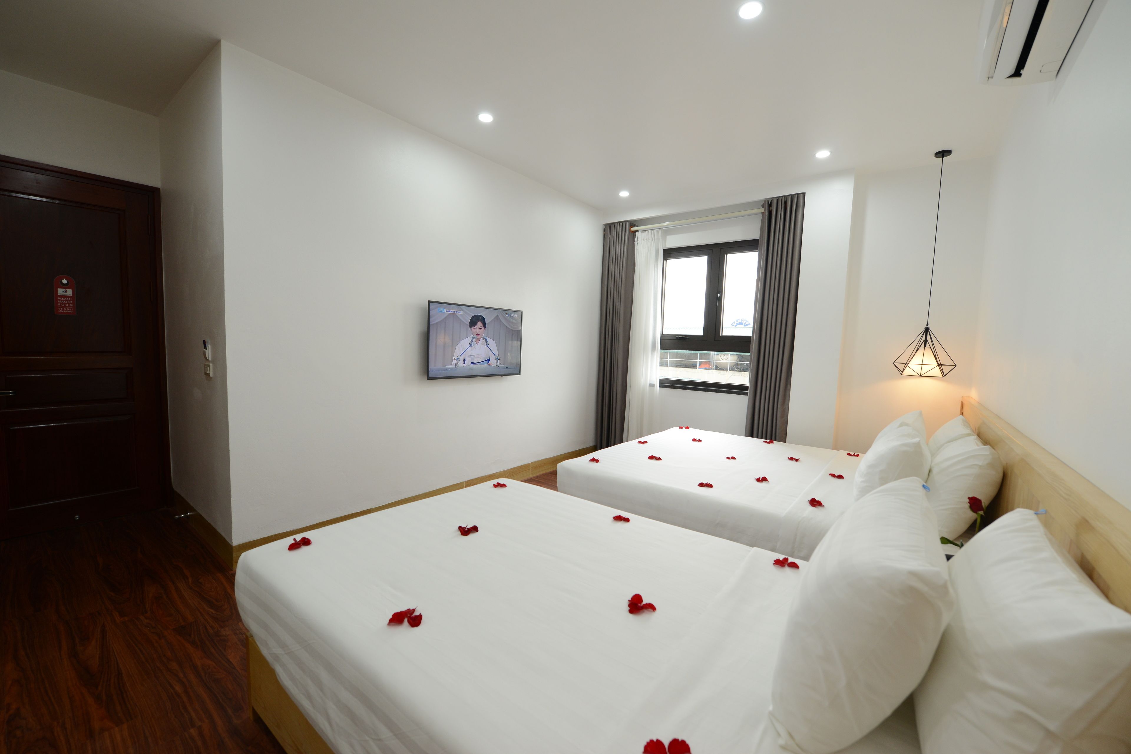 Une chambre d’hôtel avec deux lits, chacun décoré de pétales de roses rouges. Il y a une télévision sur le côté gauche, une fenêtre avec des rideaux et un luminaire suspendu.