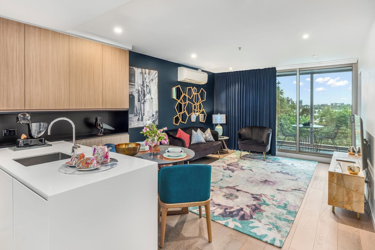 A modern apartment with a kitchen, dining area, and living room. It features a large window, a black sofa, and a blue chair.
