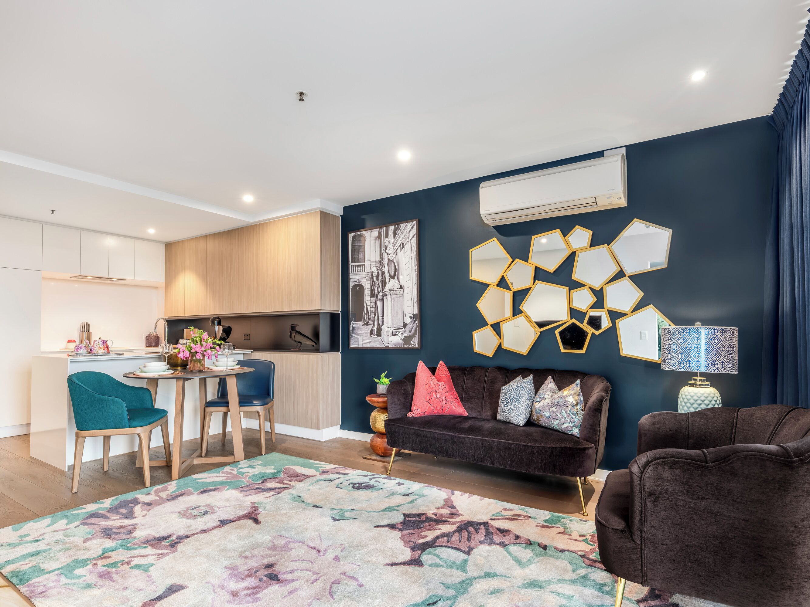 A modern living room with a dark blue accent wall, a brown sofa, teal chairs, and a floral rug.