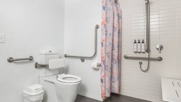A modern bathroom with a white toilet, a grab bar, a shower with a handheld showerhead, and a roll of toilet paper on the wall.