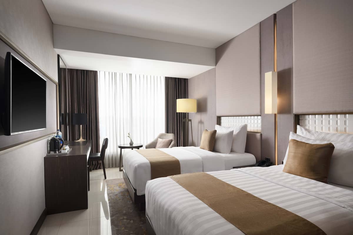 A hotel room with two beds, a desk, a chair, a TV, and a window with curtains.