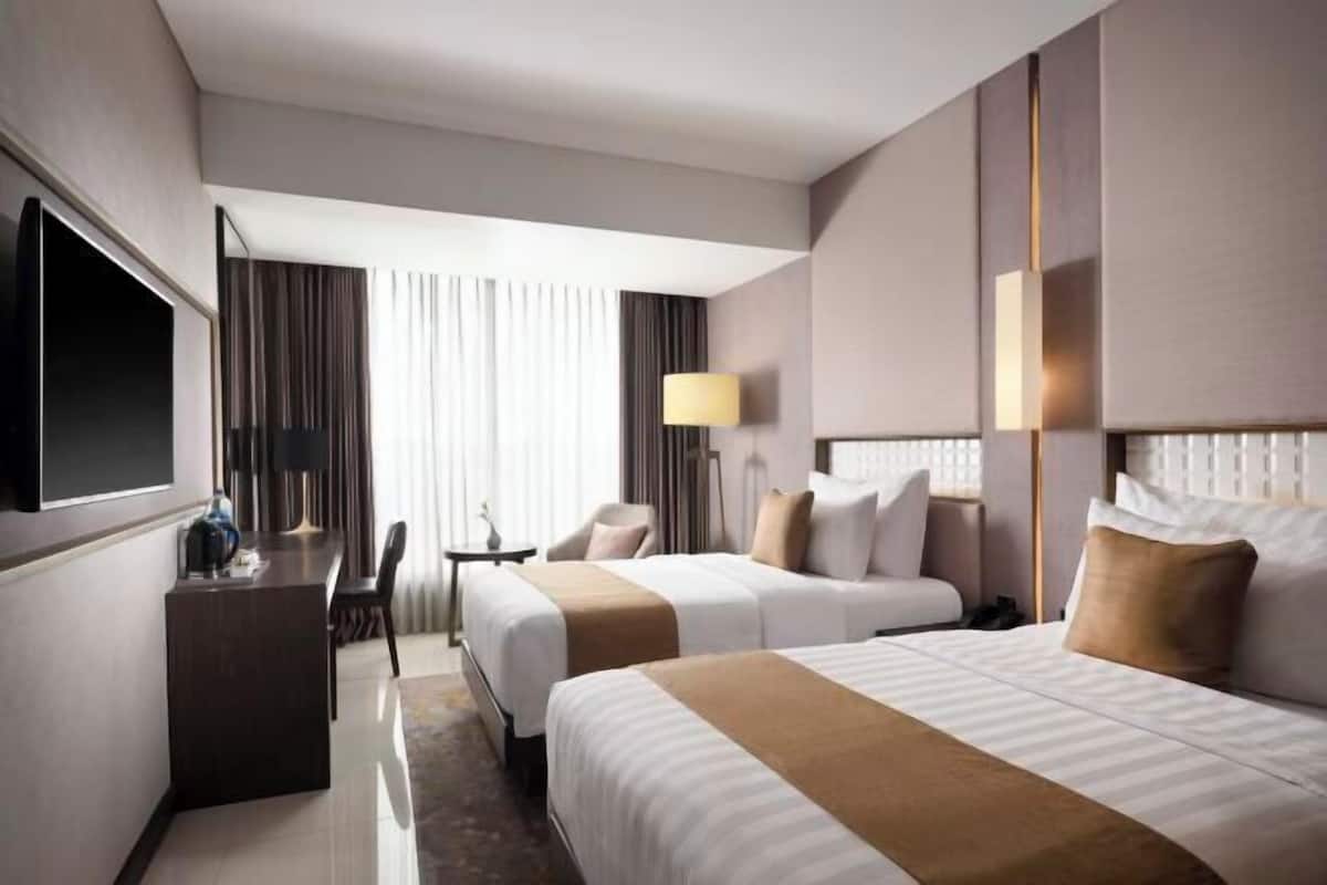 Ein Hotelzimmer mit zwei Betten, einem Schreibtisch, einem Fernseher und einem fensterbepflanzten Fenster.