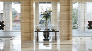 Un vestíbulo con suelo de mármol, una mesa central con un jarrón de flores y grandes ventanales con vistas a edificios y árboles.