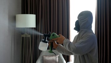 Una persona con equipo de protección usando un pulverizador en una habitación con una lámpara y cortinas.