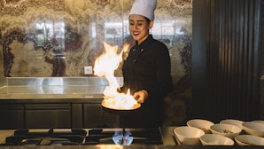 Un chef en una cocina profesional con una estufa a gas, fuego y diversos utensilios de cocina.