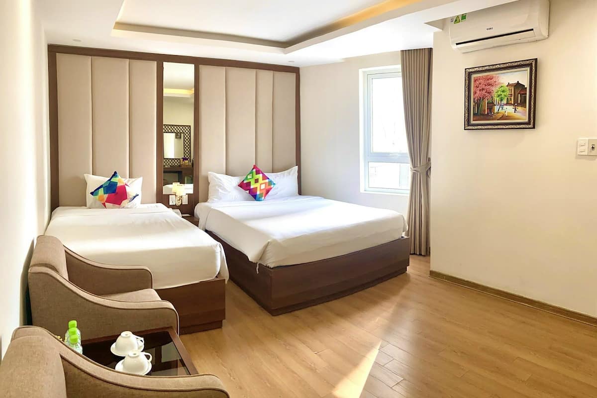 A hotel room with two beds, a sofa, a small table with a teapot and cups, an air conditioning unit, and a window with curtains.