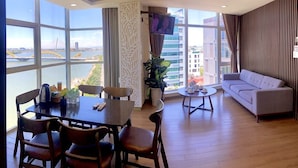 A modern living room with a dining area, large windows offering a city view, a sofa, and a TV. - Pariat River Front Hotel (Da Nang)