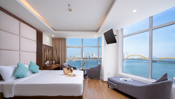 A modern hotel room with a large bed, a sofa, a TV, and a view of the city and water. - Pariat River Front Hotel (Da Nang)