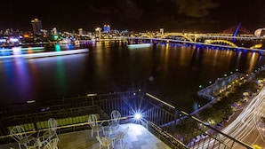 A nighttime cityscape with a river, illuminated bridge, and a view from an elevated balcony with chairs. - Pariat River Front Hotel (Da Nang)