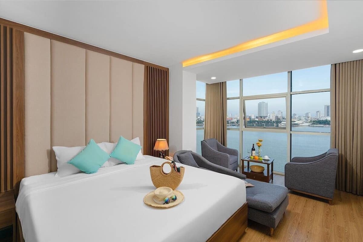 A hotel room with a bed, a view of the city, a table with a lamp, and a chair.