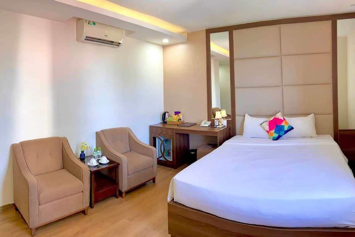 A hotel room with a bed, two armchairs, a desk with a mirror, and an air conditioning unit.
