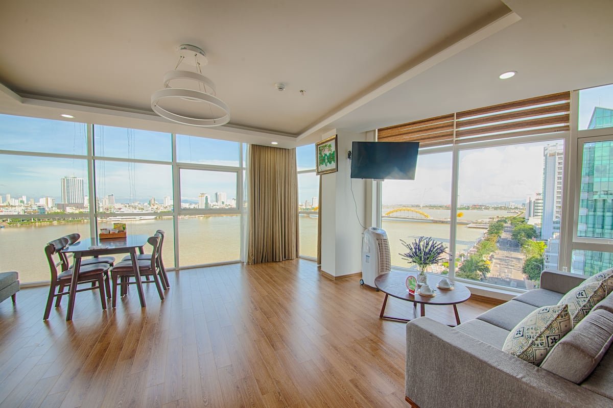 A spacious living room with a large window offering a city view, a flat-screen TV, a dining area with chairs, and a sofa.