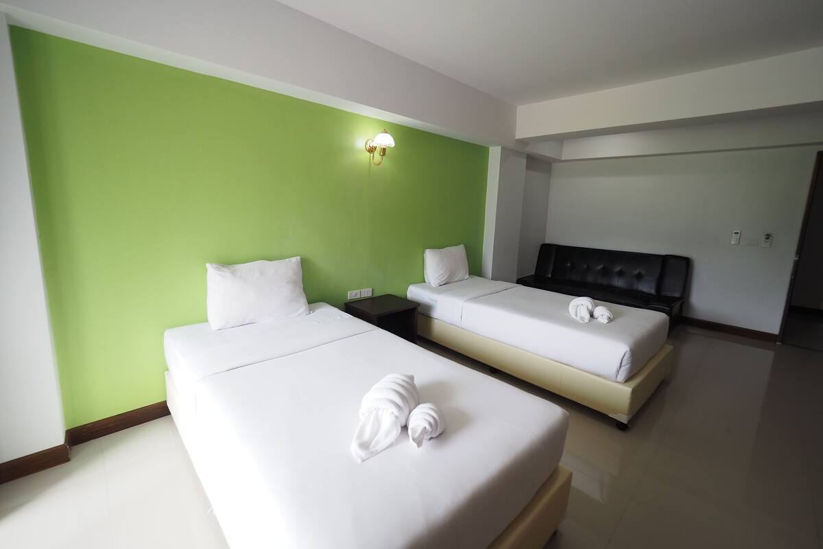 A hotel room with two beds, green walls, white bedding, a nightstand, and a wall-mounted light fixture.