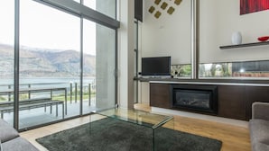 A modern living room with a glass coffee table, a flat-screen TV, a fireplace, and a scenic view of a lake and mountains. - Lakeshore Springs Apartments (Wanaka)